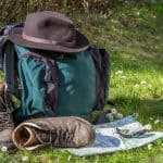 A bug out bag pictured with a hat, boots, and a map in a grassy field.