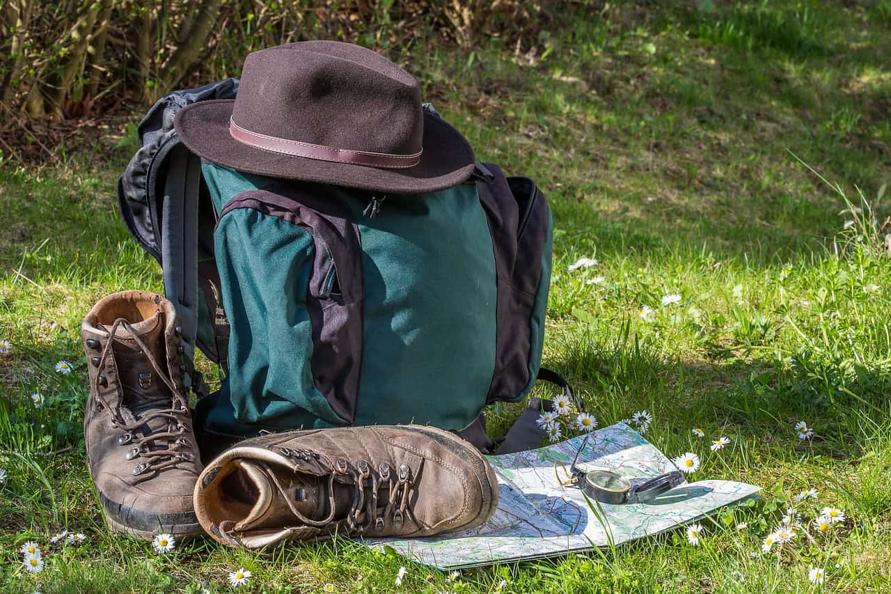 A bug out bag pictured with a hat, boots, and a map in a grassy field.