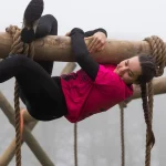 A woman climbing upside down along a wooden beam in a survival run course, representing Basic Skills Every Prepper Should Learn