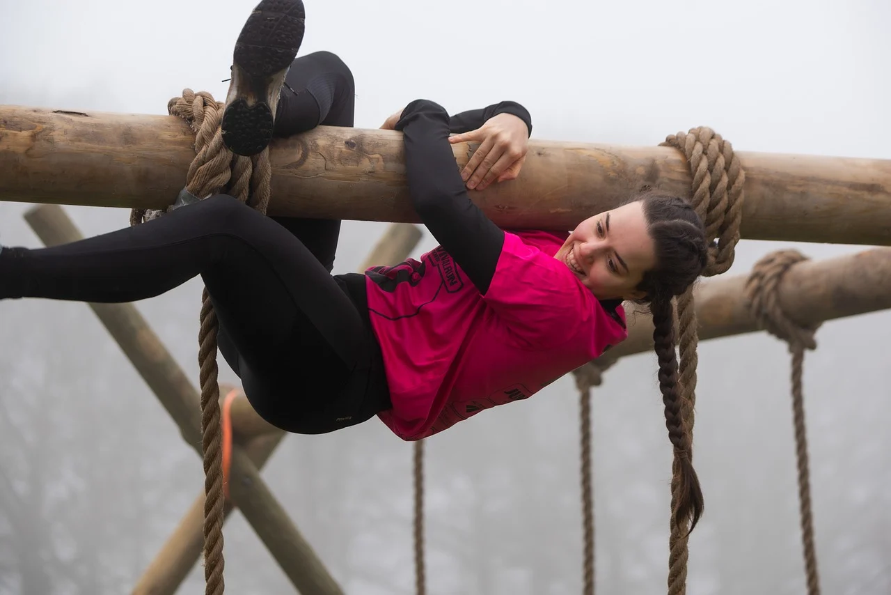 A woman climbing upside down along a wooden beam in a survival run course, representing Basic Skills Every Prepper Should Learn