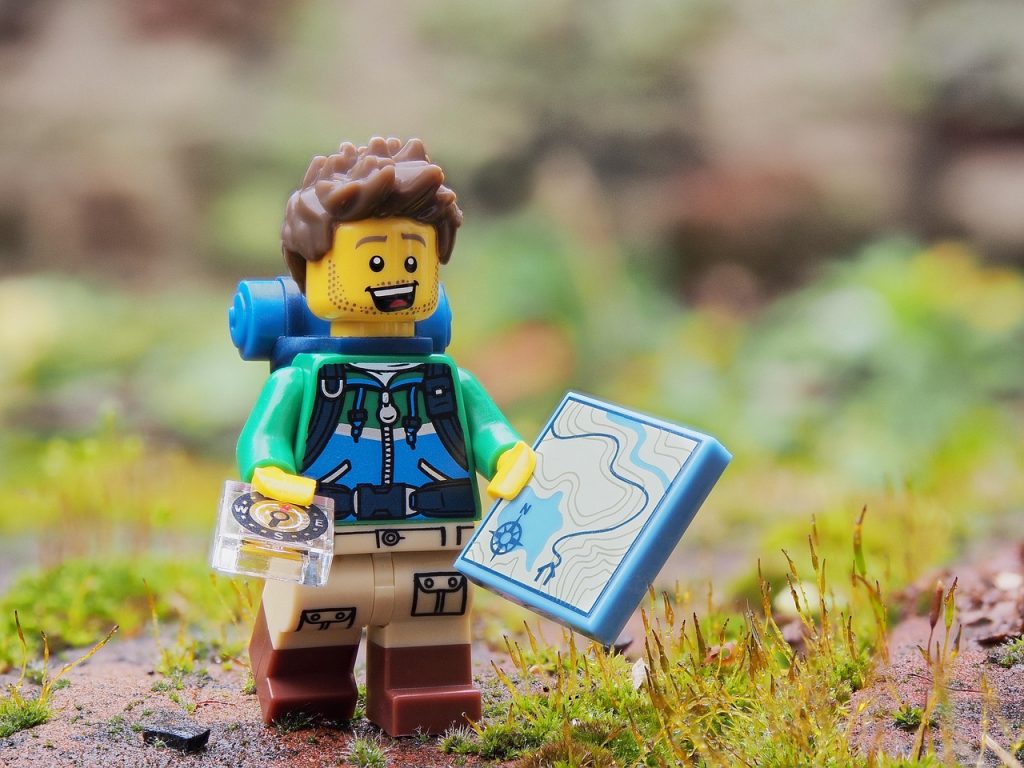 A brick figurine in a hiking outfit with a backpack on, holding a map and compass, representing "What to Include in Your First Bug Out Bag"